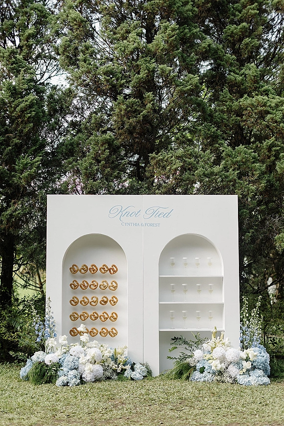 Wedding welcome sign on a white arched backdrop with calligraphy, hydrangea florals, champagne flutes and pretzels on a lawn by evergreens