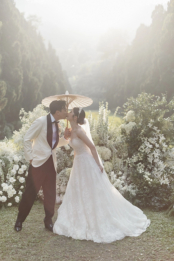 Wedding kiss portrait of bride and groom kissing under a paper parasol, lace gown and white tuxedo, backlit on a garden lawn with hills
