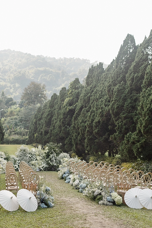 Ceremony aisle design with floral aisle arrangements of hydrangeas lining wooden chairs on a lawn, with mountains in the distance