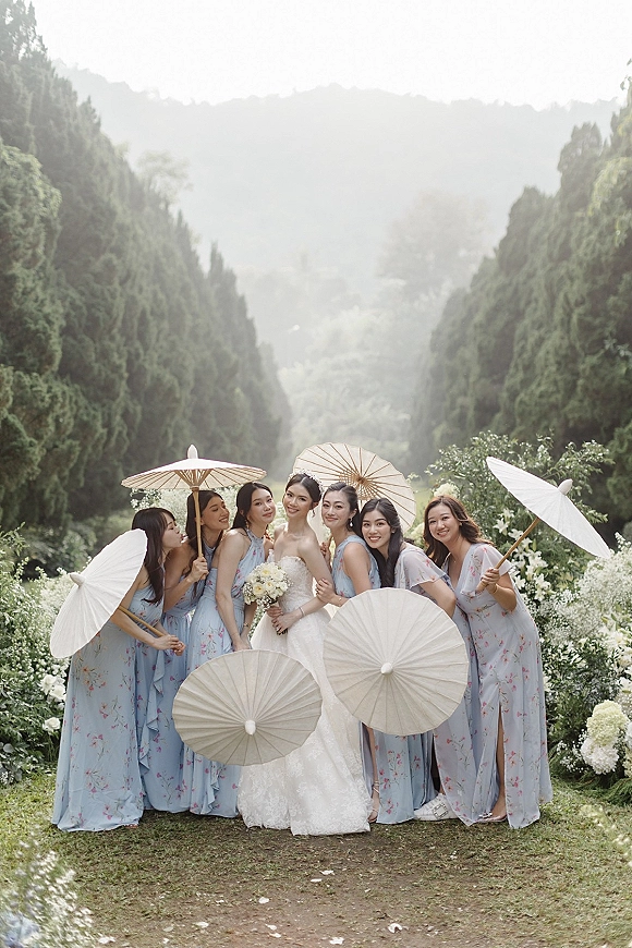 Bride with bridesmaids holding paper parasols, in blue floral dresses beside her lace gown on a garden path with mountain backdrop
