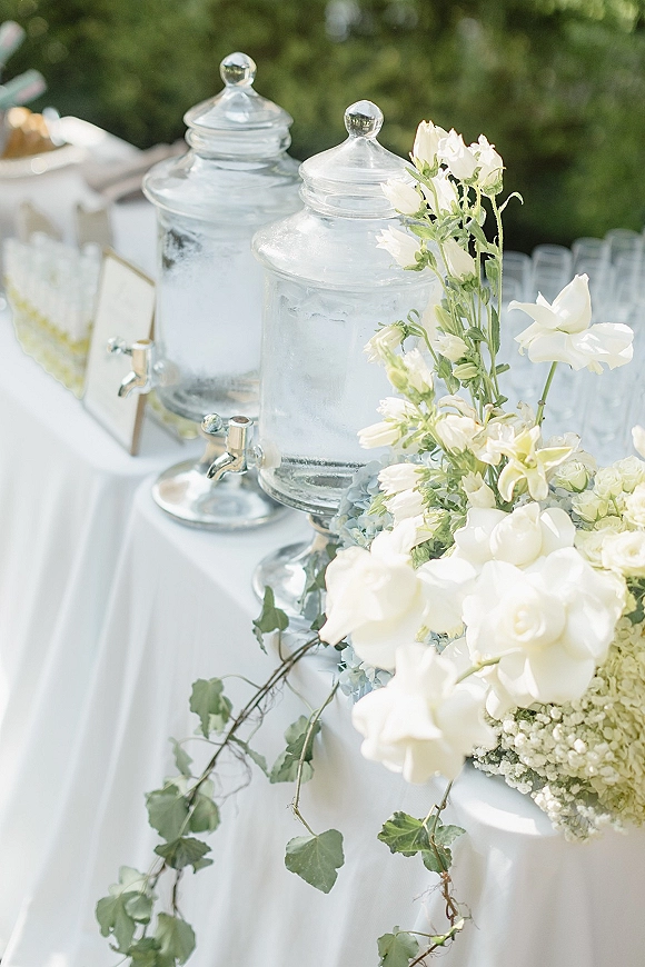 Drink dispenser station with glass beverage dispensers and metal spigots, styled with white roses and greenery garland outdoors among trees