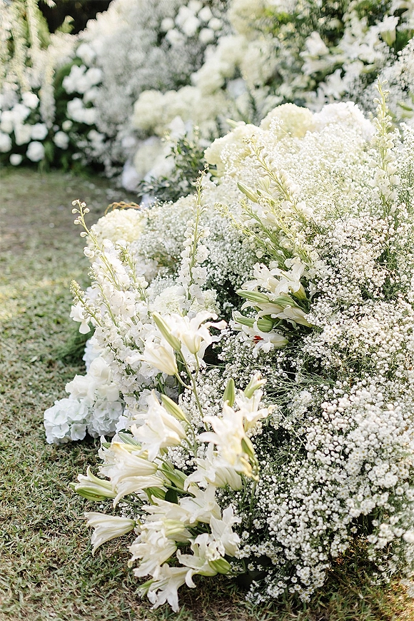 Ceremony aisle florals with white lilies and baby's breath in ground arrangements lining a grass lawn aisle amid garden greenery