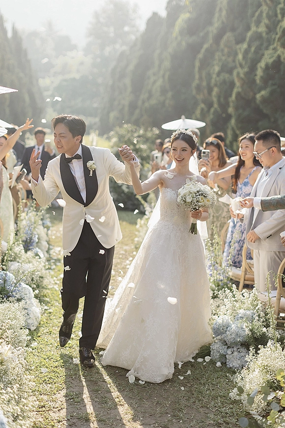 Wedding recessional as bride and groom walk down a garden aisle, guests tossing flower petals, bride in tiara and veil holding bouquet