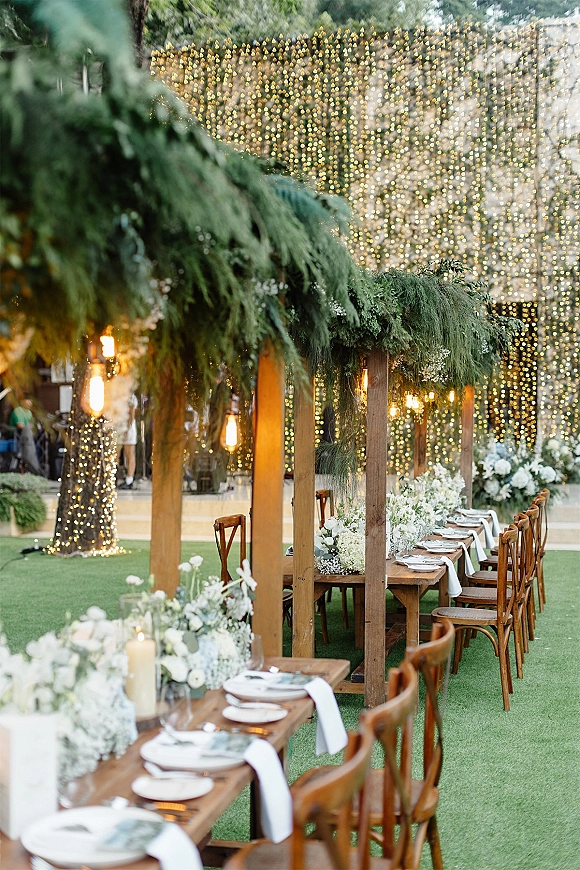 Reception tablescape at an outdoor wedding reception with a long farm table, greenery garland, white flowers, candles, and string lights above trees