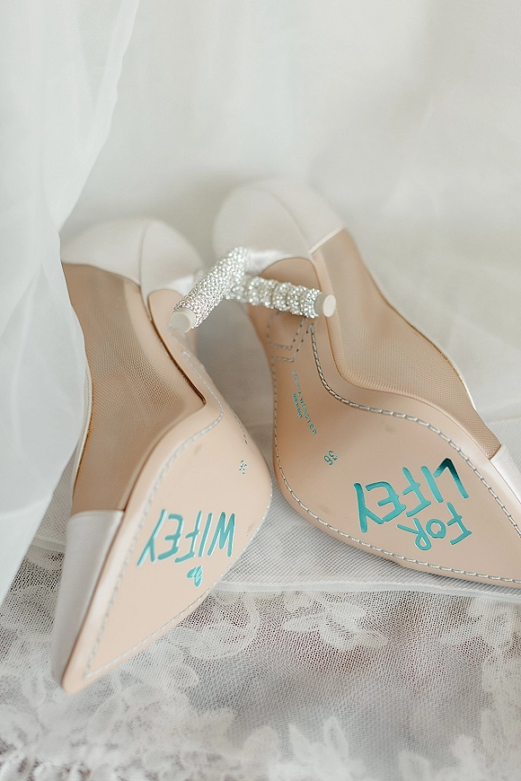 Bridal shoes with white pointed-toe heels and a crystal ankle strap, shown on white lace fabric with sole lettering visible