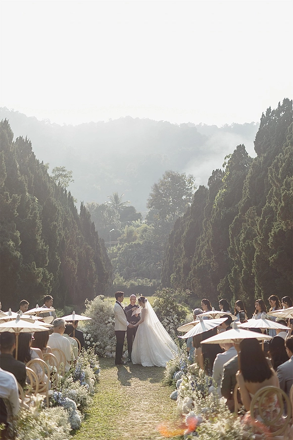 Outdoor wedding ceremony with bride and groom at the altar beneath a flowered arch, floral-lined aisle, guests with parasols on a garden lawn