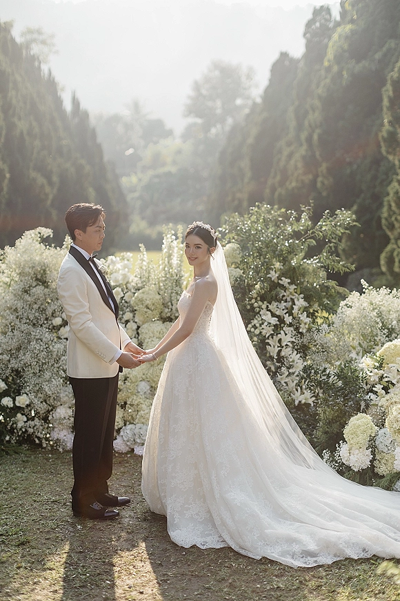 Couple portrait of bride and groom holding hands under a white floral arch, her long veil and tiara glowing in sunlit garden.