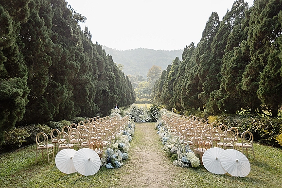 Outdoor ceremony setup with wood chairs and a floral lined ceremony aisle of hydrangeas, baby's breath, and white paper parasols on a grassy lawn