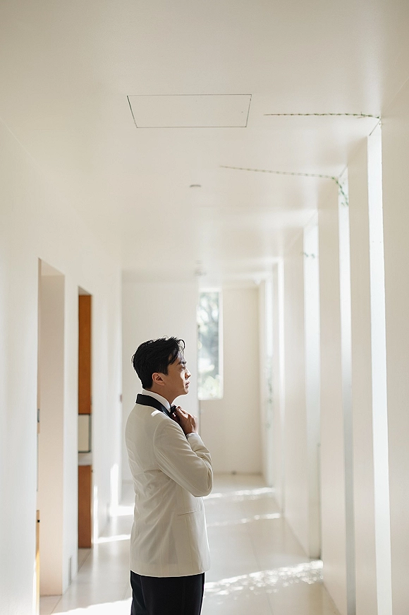 Groom portrait with the groom adjusting bow tie in a white tuxedo jacket, standing in a bright sunlit hallway with columns