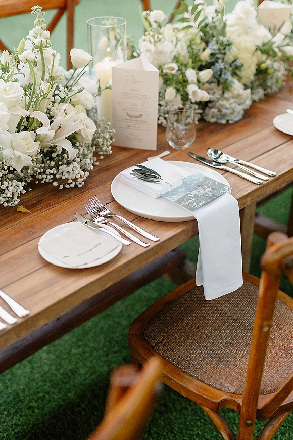Reception tablescape on a farmhouse wedding table with white rose and lily centerpiece, pillar candles in hurricanes, and place cards on a green lawn