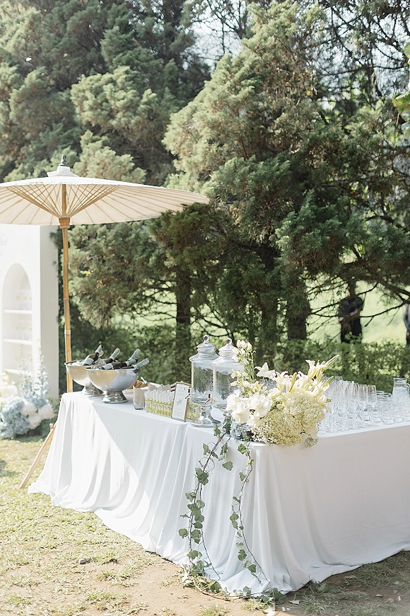 Wedding bar setup with glass beverage dispensers, champagne bottles in ice buckets, and white linens under an umbrella on a lawn near a tent