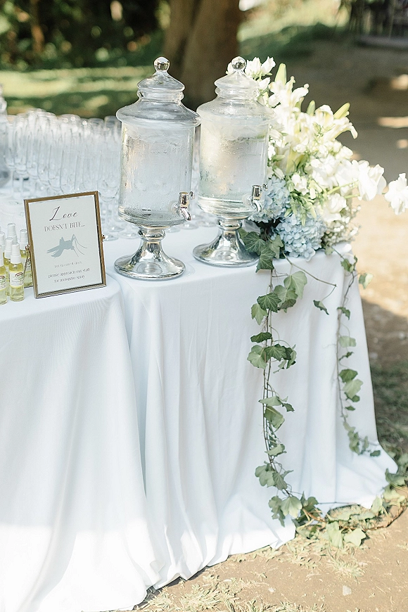 Wedding drink station with glass water dispensers, drinking glasses, and hydrangea greenery on a white tablecloth, set on a sunny lawn with trees