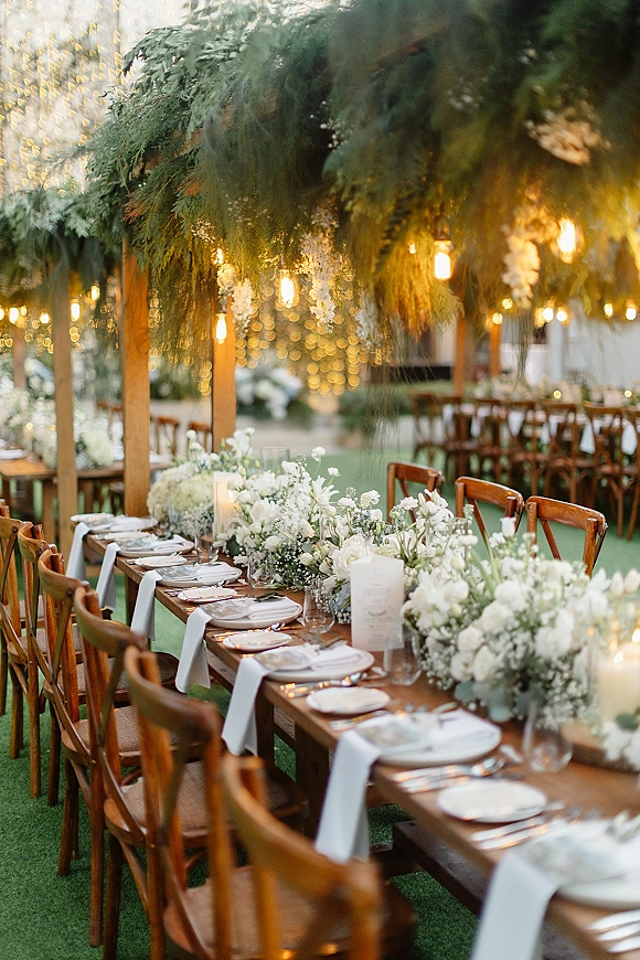 Reception tablescape with white floral centerpieces and greenery garland on wood farm tables, candlelight and string lights in a garden lawn setting