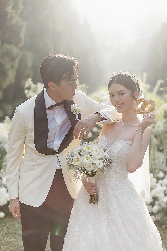 Couple portrait of bride in lace dress and veil holding a bouquet beside groom in a white tuxedo jacket, with sunlit garden trees behind