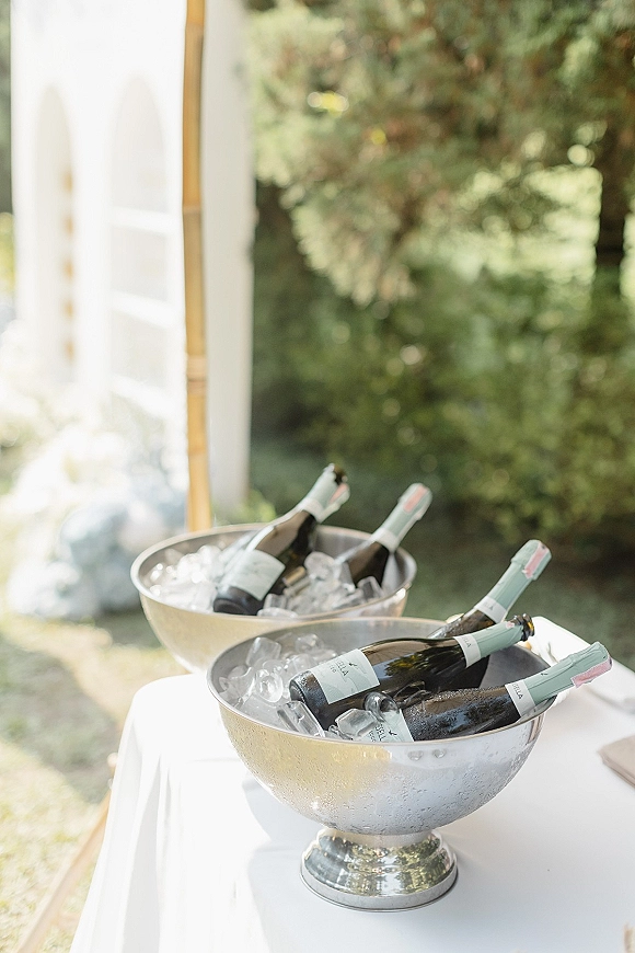 Champagne bucket display with wedding champagne station bottles on ice in silver tubs on a white tablecloth amid lush greenery outdoors