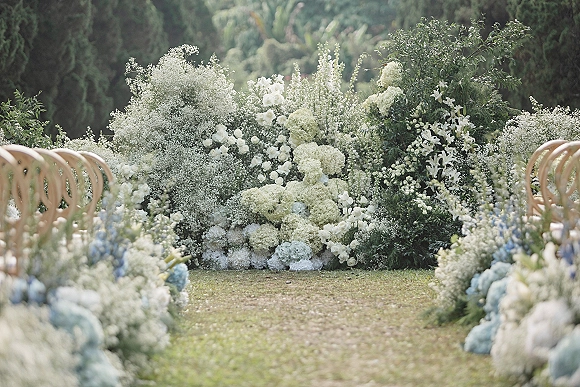Ceremony floral backdrop of white roses, hydrangeas and baby's breath with greenery, set beside wooden chairs on a garden grass aisle