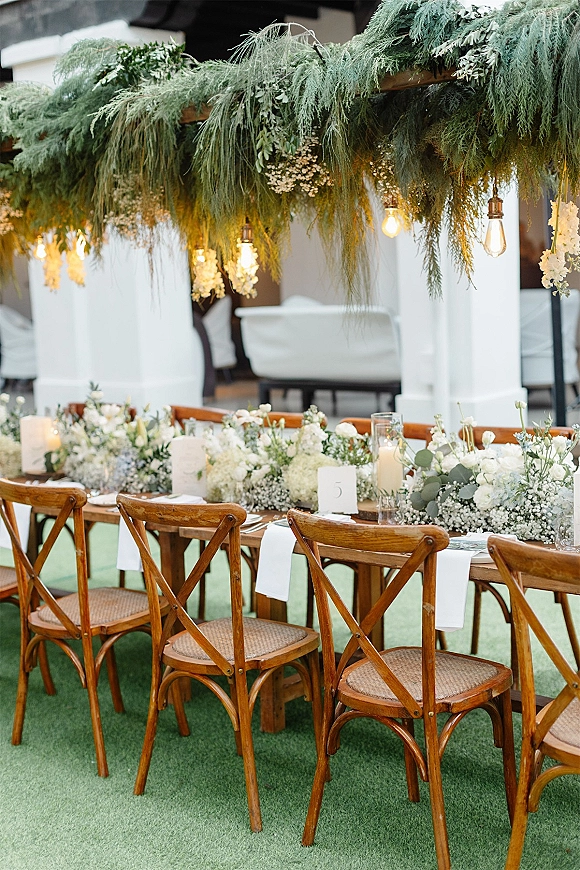 Reception tablescape with farmhouse wedding table, white runner, white florals and greenery garland, taper candles, Edison bulbs on patio by lawn