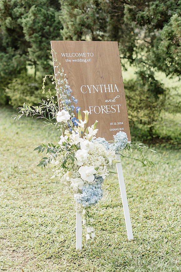 Wedding welcome sign with blue hydrangea and white flowers on a white stand, set on a grass lawn with evergreen trees behind