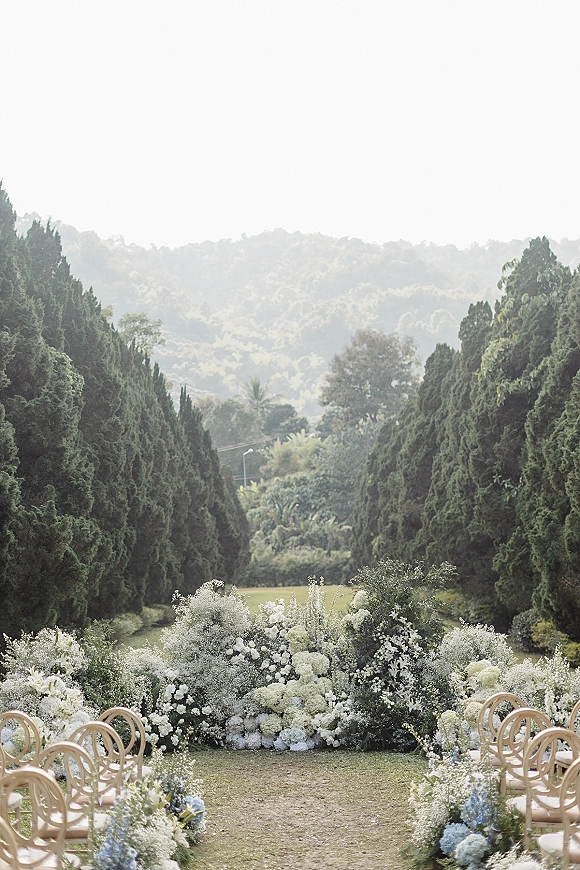 Outdoor ceremony setup with garden wedding ceremony aisle flowers and a floral altar arrangement on a lawn framed by tall evergreens and hills