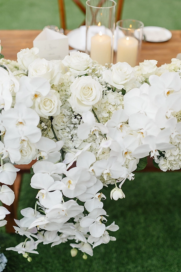 Wedding tablescape with a white floral centerpiece of orchids, roses, and hydrangea, pillar candles in glass hurricanes on a wooden table set on a green lawn
