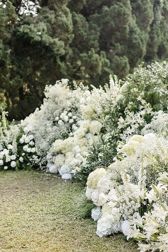 Wedding aisle flowers forming a floral aisle runner with white hydrangeas and baby's breath along a grassy garden path amid evergreens