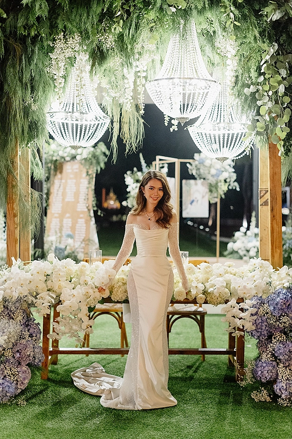 Bridal portrait of a bride in an off the shoulder wedding dress with lace gloves, seated beneath a crystal chandelier and floral arch in a nighttime garden