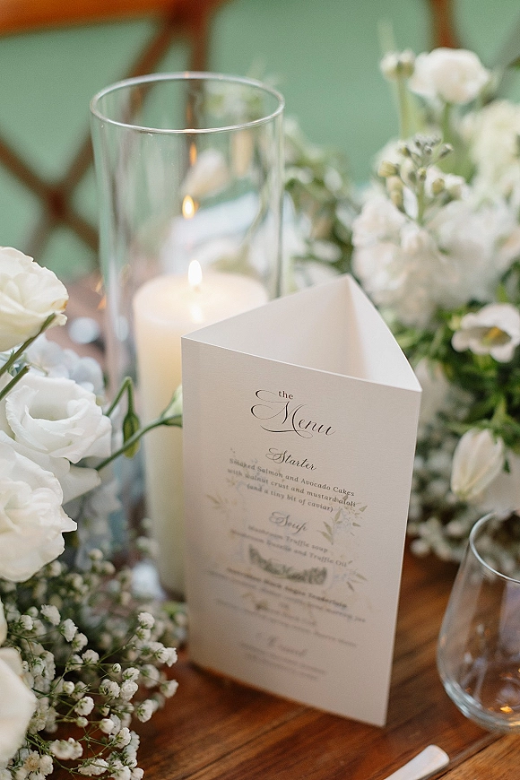 Wedding menu card with calligraphy on a folded wedding menu beside white flowers, baby’s breath and a pillar candle on a wood table