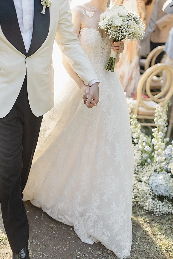 Wedding recessional bride and groom holding hands, walking down a sunlit outdoor aisle as guests toss flower petals, bouquet in hand