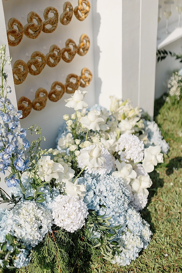 Wedding pretzel wall with hanging pretzels and blue hydrangea floral accents against a white wall on lawn grass outdoors