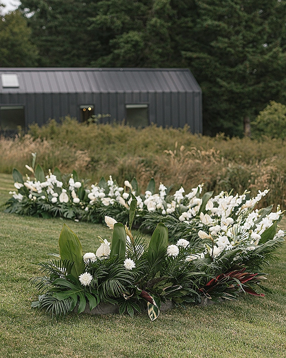 Wedding ceremony flowers form a ceremony floral meadow of white blooms and tropical greenery along a grass lawn beside a metal barn building