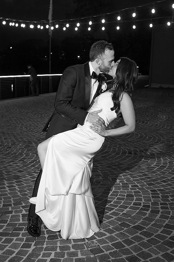 Wedding kiss portrait of bride in a strapless satin dress and groom in tuxedo dipping under string lights on a cobblestone patio at night