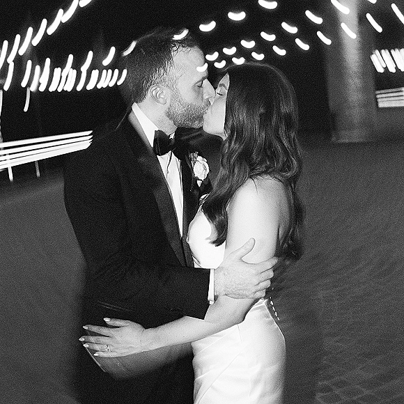 Wedding kiss portrait of newlyweds kissing, bride in bridal gown showing engagement ring, groom in tuxedo with boutonniere under string lights at night