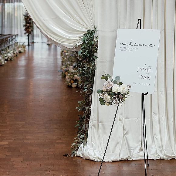 Wedding welcome sign on easel with greenery garland and floral accent, set before a white curtain backdrop with aisle florals and chairs