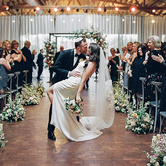 Wedding kiss moment as groom dips bride for a kiss, her long veil and bouquet flowing down an indoor aisle with string lights overhead