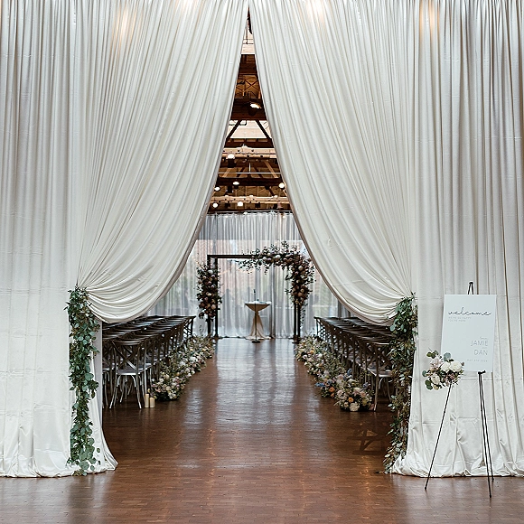 Ceremony setup for an indoor wedding ceremony with white fabric draping, floral-lined aisle, and a greenery arch under string lights and beams