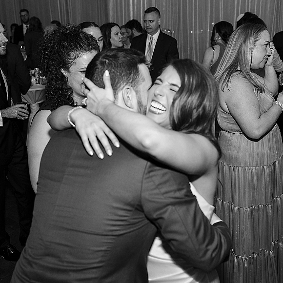 Wedding reception moment as bride hugs groom while he kisses her cheek, cocktail glasses raised by guests amid draped curtains indoors
