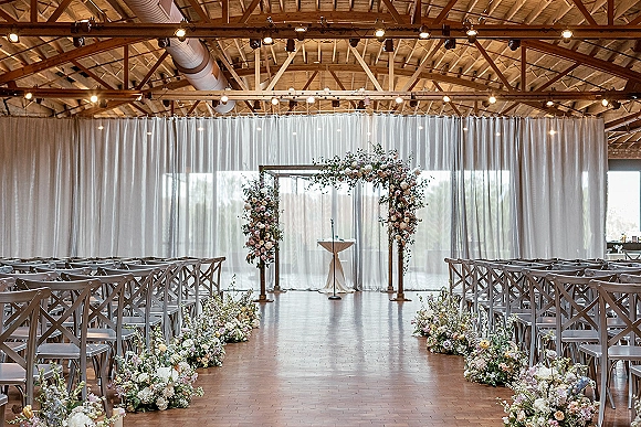 Ceremony setup with floral arch and aisle florals, white draping and string lights in a barn interior with wood beams and large windows