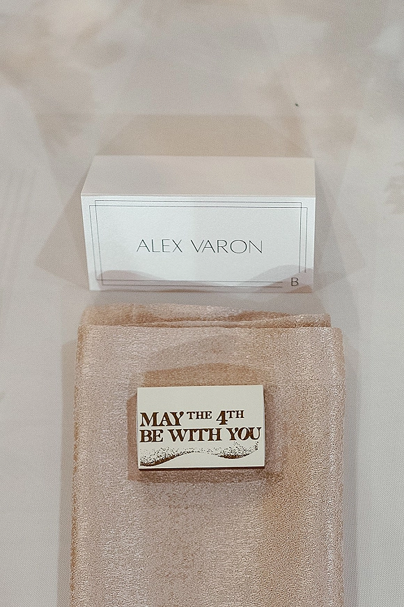Wedding place card with minimalist place card typography resting on a beige napkin, featuring a small favor tag on the table surface