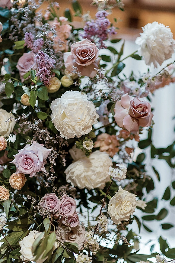Wedding floral arrangement with pastel roses and peonies, finished with lilac accents and airy greenery against blurred indoor architecture