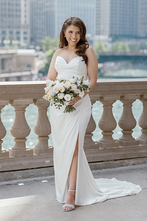 Bridal portrait of a bride holding bouquet in a strapless satin dress with slit on a stone terrace by a river and city skyline