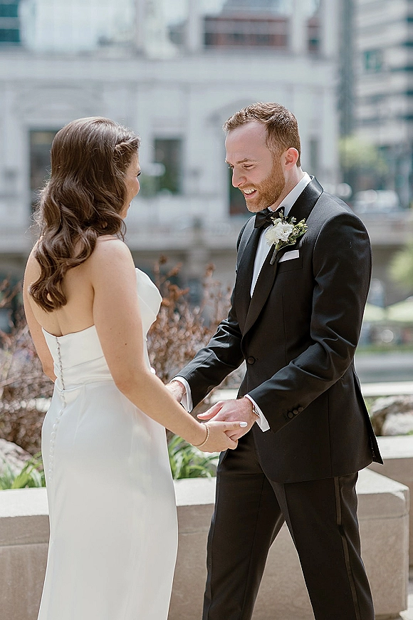 First look moment as bride in a strapless wedding dress holds hands with groom in black tuxedo, smiling in an urban courtyard