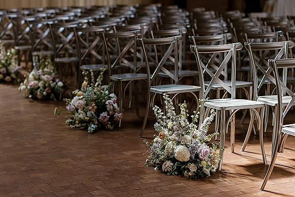 Ceremony aisle decor with aisle florals ground arrangements of pastel and white blooms with greenery beside metal cross-back chairs on a parquet floor
