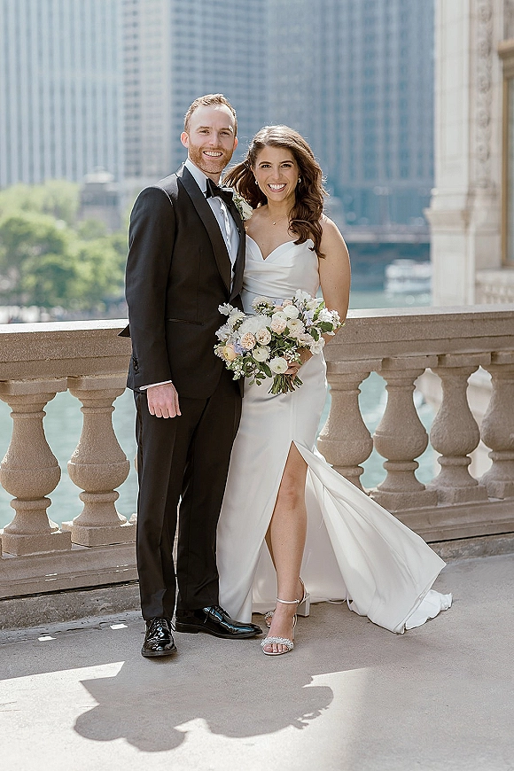Couple portrait of bride holding a white rose bouquet beside groom in black tuxedo, posing by a riverfront balustrade with skyline behind