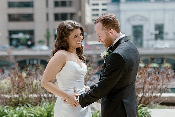 Couple portrait of bride and groom holding hands, bride in strapless dress and groom in tuxedo, with city greenery backdrop