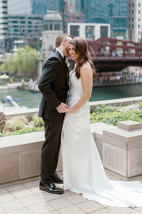 Couple portrait of bride and groom holding hands as he whispers to her on a stone riverwalk terrace with skyline and bridge behind
