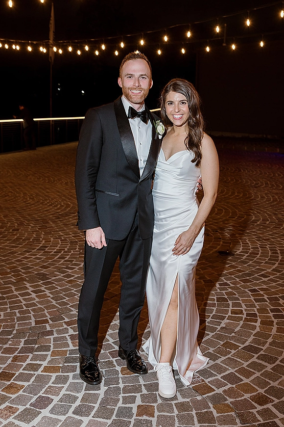 Couple portrait of bride in a slit satin dress and sneakers with groom in black tuxedo under string lights on a cobblestone patio at night