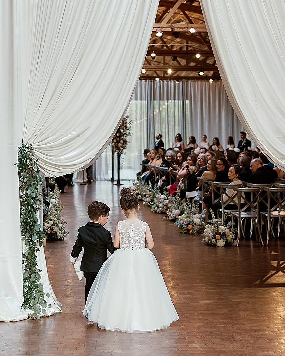 Ceremony processional with flower girl and ring bearer walking down a flower-lined aisle under string lights in a rustic barn venue