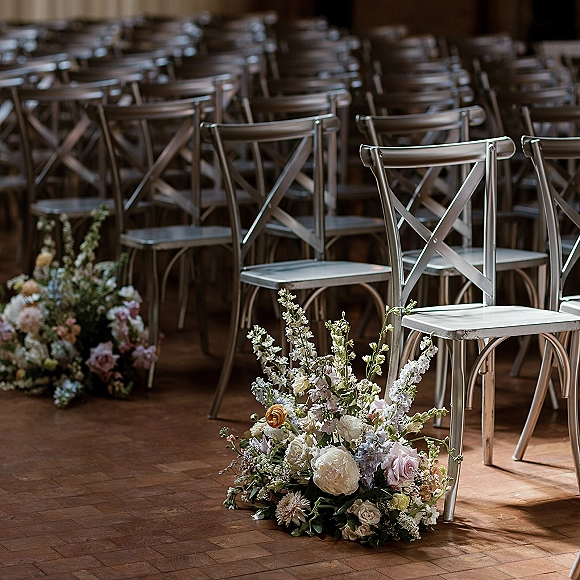 Ceremony seating with wedding aisle chairs in neat rows, metal cross back chairs and soft pastel aisle florals on a wood floor indoors