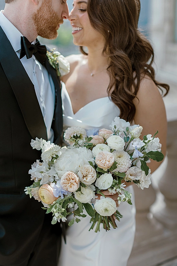 Couple portrait of bride and groom close up, bride holding bouquet with necklace and tuxedo, by stone columns and greenery outdoors