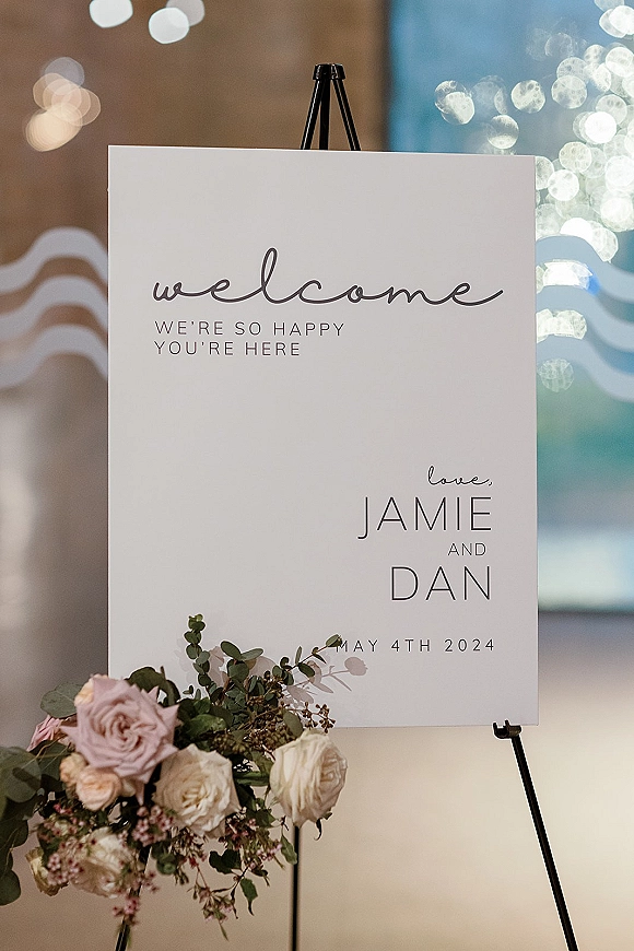 Wedding welcome sign on a black easel with rose and eucalyptus floral corner accent, set against a softly blurred indoor bokeh background